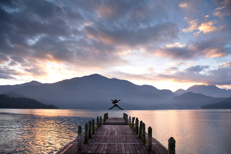 Person jumping off a dock into a lake with mountains and a colorful sky.