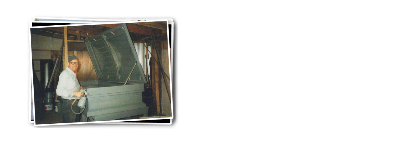 Vintage Photo of Forrest Davis smiling at the camera and standing in front of a metal oversized casket with a spray paint machine ready to paint the casket in a barn